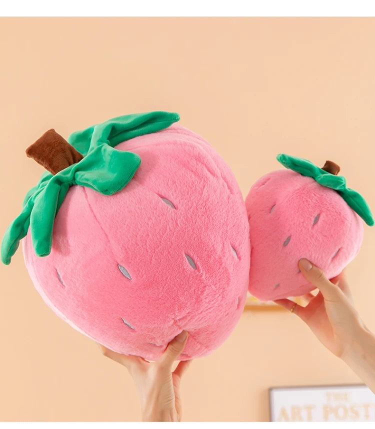 A pink strawberry plush throw pillow with green leaves and a brown stem being held by hands.