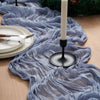 A gray crinkle gauze table runner draped over a wooden surface with a white candle and greenery.