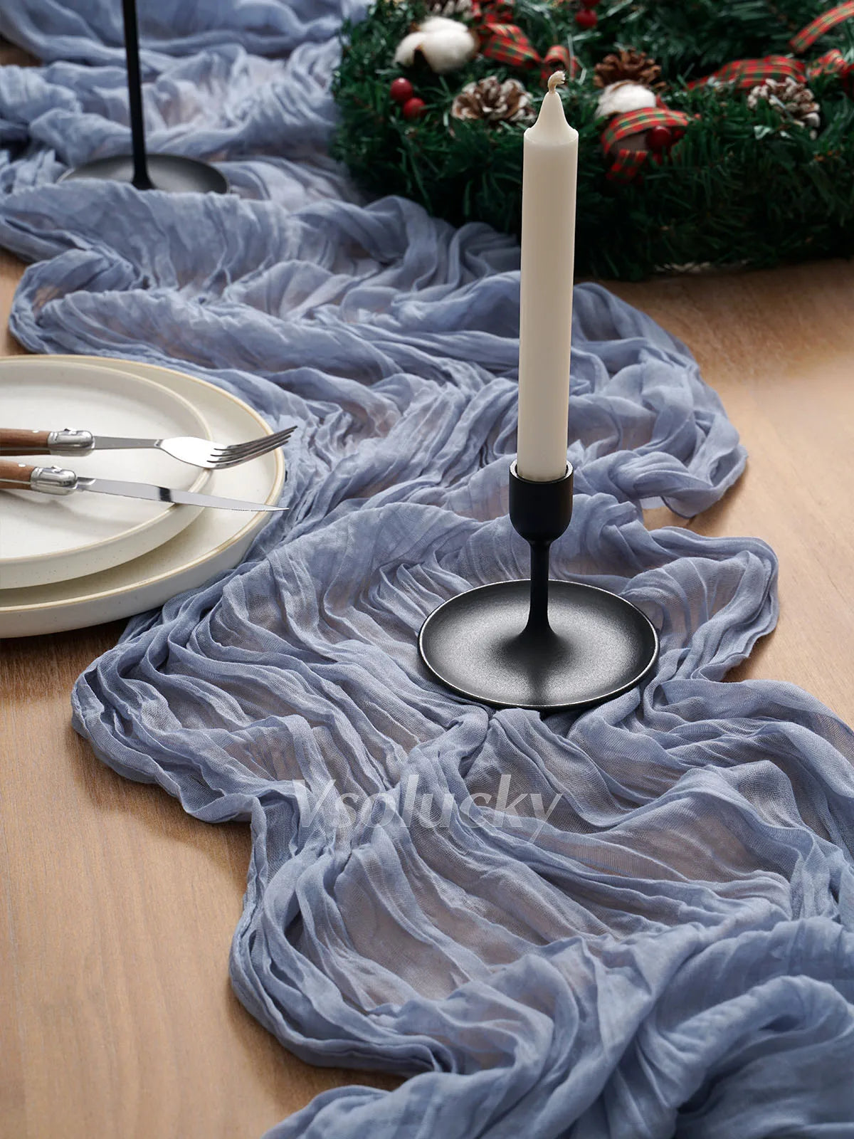 A gray crinkle gauze table runner draped over a wooden surface with a white candle and greenery.