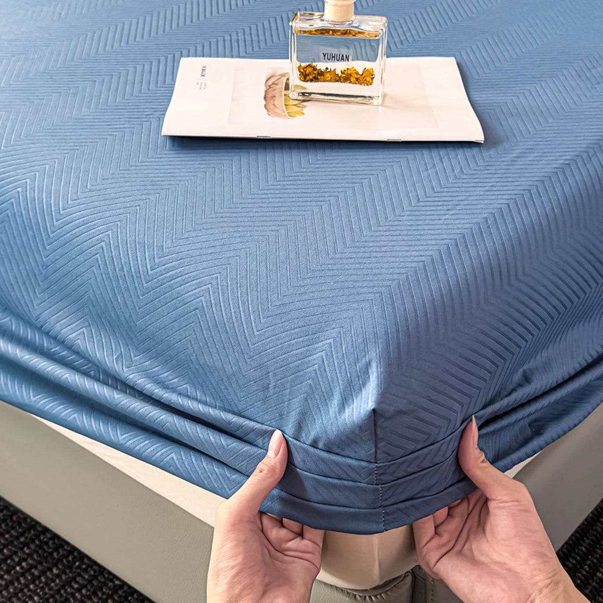 A blue fitted sheet is being held up, showing a small glass bottle of YUMIAN oil and a piece of dried plant material on the s
