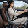 A woman is sitting in a car, driving, wearing a beige shirt and pants, with an ergonomic seat cushion visible.