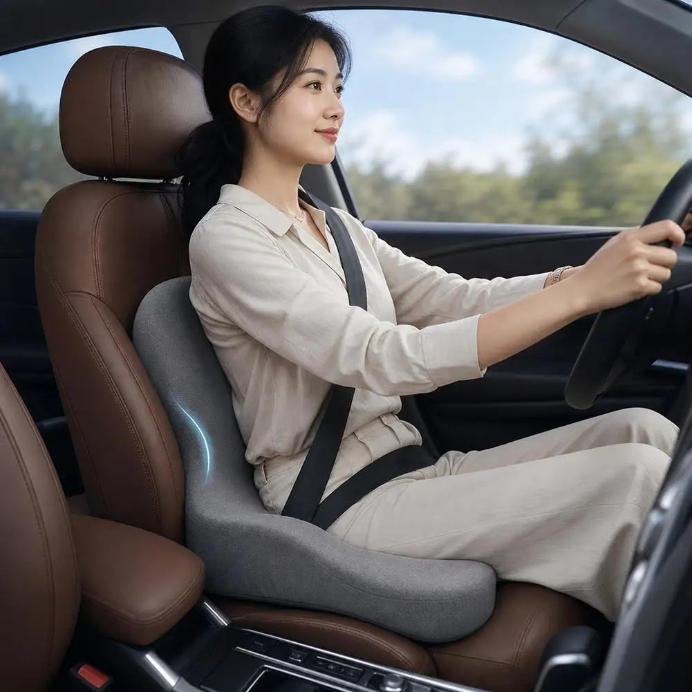 A woman is sitting in a car, driving, wearing a beige shirt and pants, with an ergonomic seat cushion visible.
