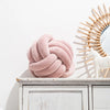 A pink knitted velvet throw pillow resting on a white wooden shelf next to a sunburst mirror.