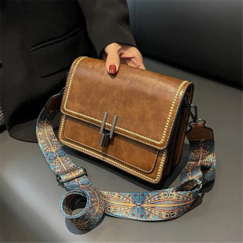 A brown leather mini cross-body bag with a patterned strap is held by a person.