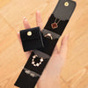 A hand holds a black PU leather jewelry organizer pouch with four compartments, featuring gold-toned jewelry.