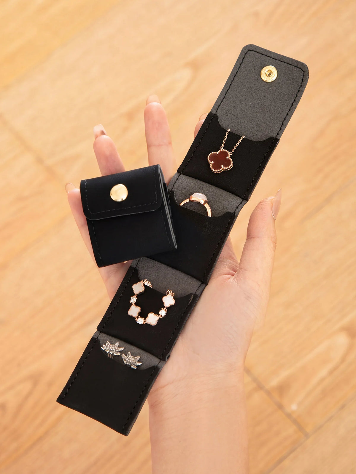A hand holds a black PU leather jewelry organizer pouch with four compartments, featuring gold-toned jewelry.