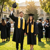 Two graduates in black academic gowns and caps stand outdoors on a sunny day.