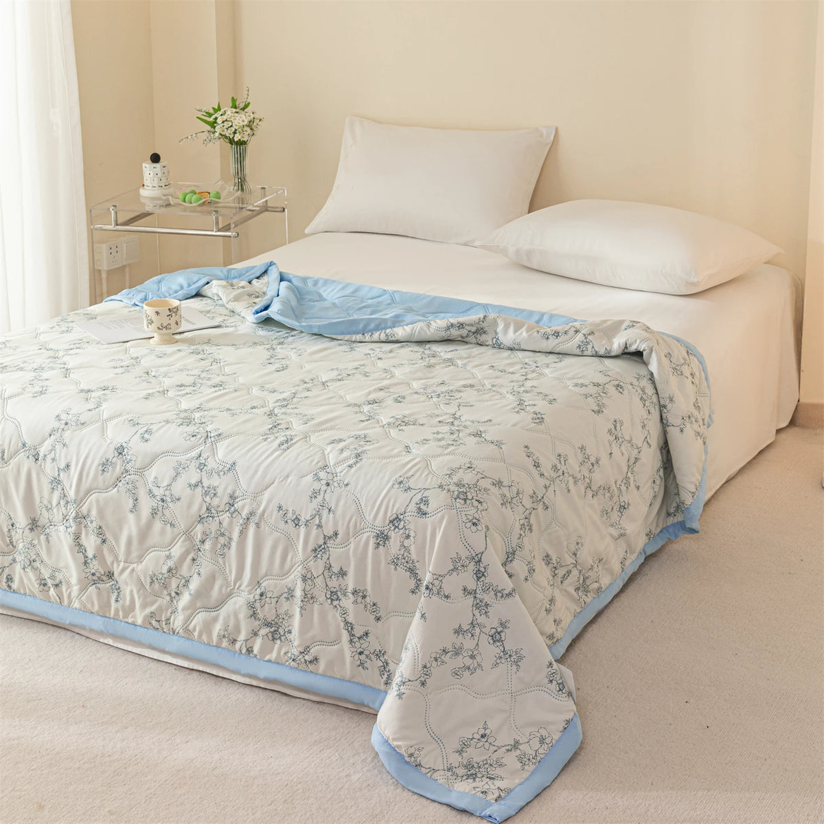 A white bed with a light blue quilt and pillows in a bright, neutral-toned bedroom.