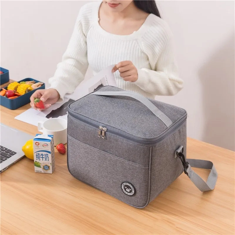 A woman holds a gray thermal insulated lunch bag with an adjustable strap on a wooden table.