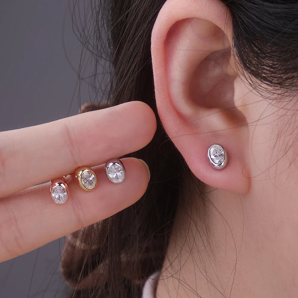 A hand is holding a pair of oval moissanite stud earrings in sterling silver near a person's ear.