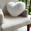 A fluffy white heart-shaped pillow resting on a light beige armchair with a wooden railing and greenery in the background.