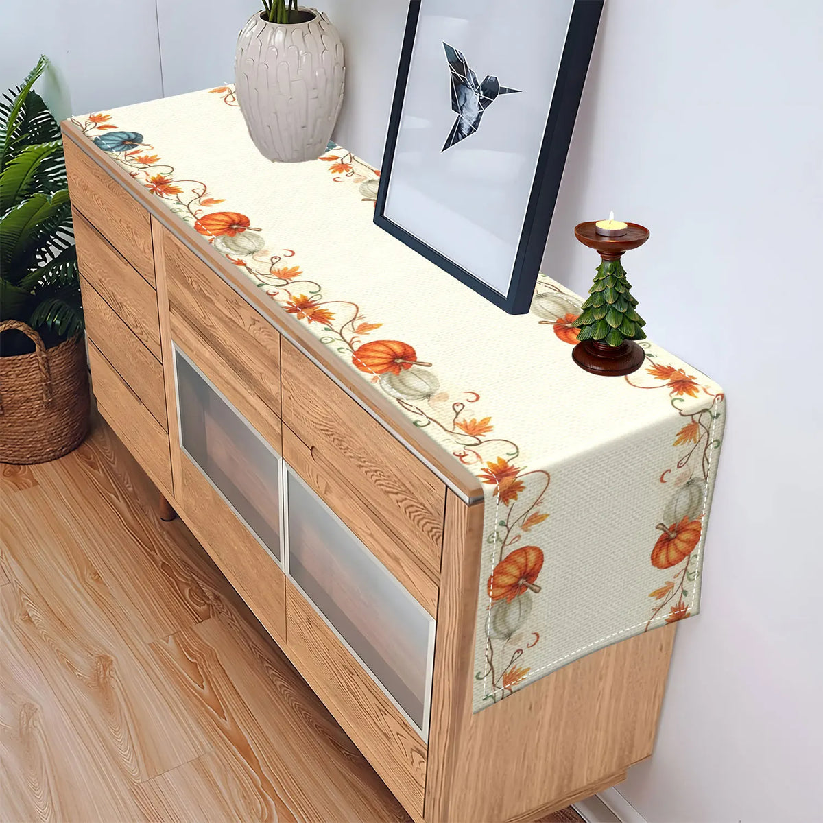 A wooden buffet cabinet decorated with a fall harvest table runner featuring orange pumpkins and autumn leaves.