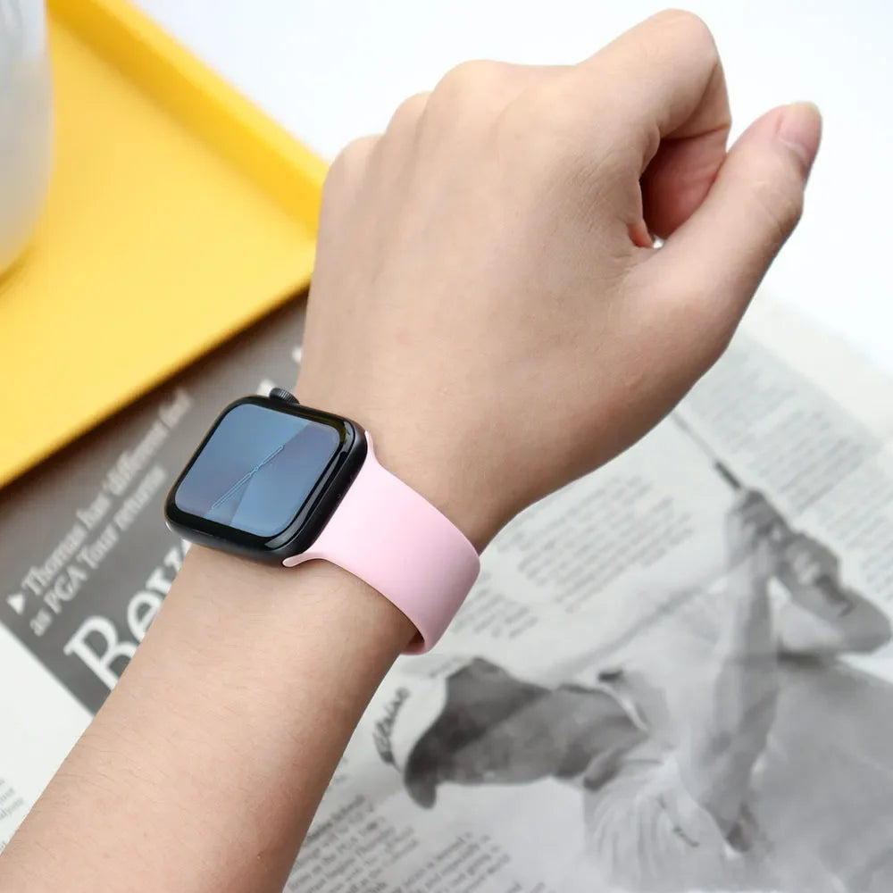 A hand wearing a pink soft silicone sport band on an Apple Watch, next to a yellow box and printed paper.