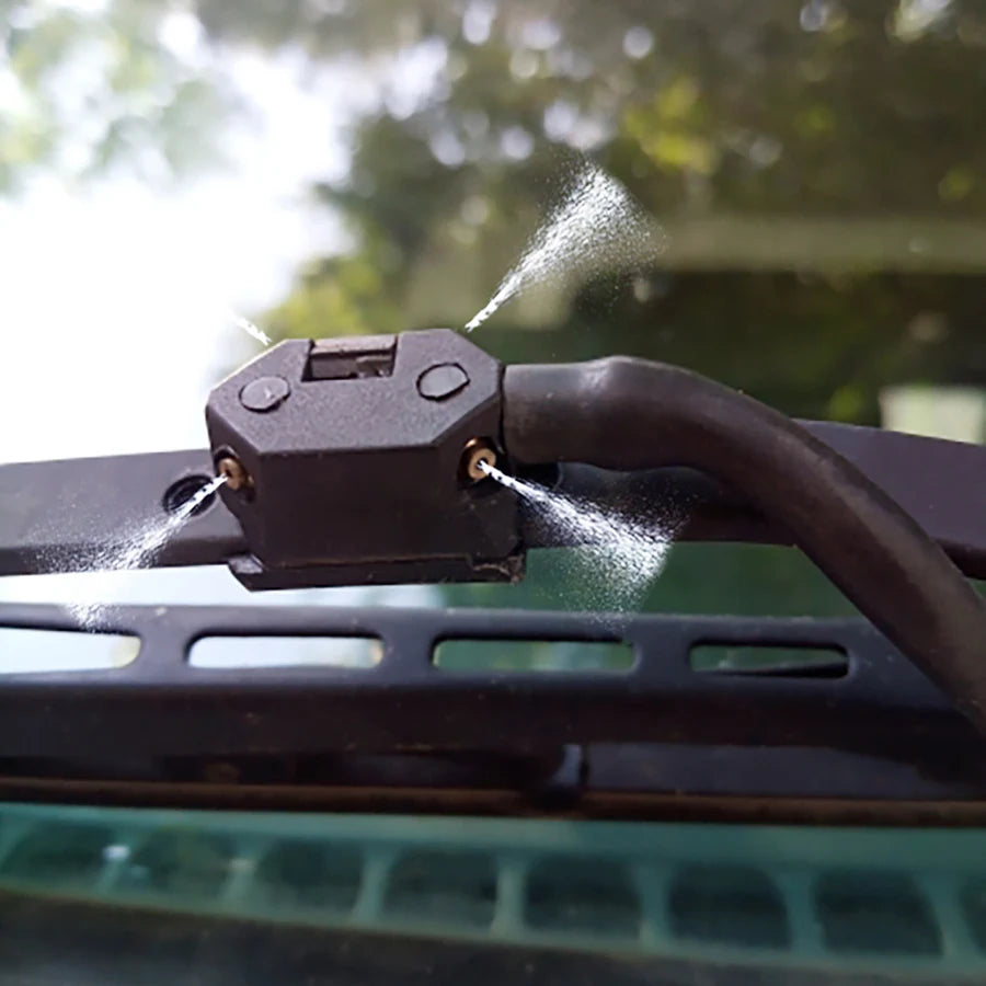 A close-up of two car windshield washer nozzles mounted on a black bracket, spraying water.