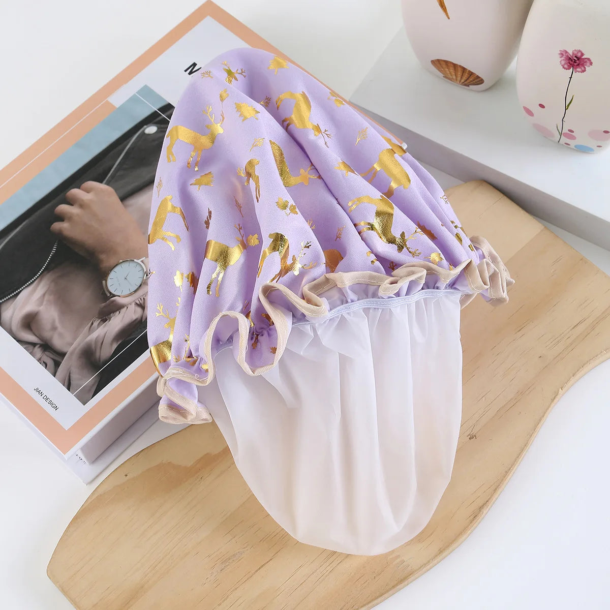 A purple and gold reusable elastic shower cap with gathered fabric and sheer lining displayed on a wooden board.
