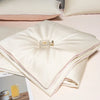 A stack of cream-colored ice silk bedding, including a quilt and pillows, resting on a light pink surface.