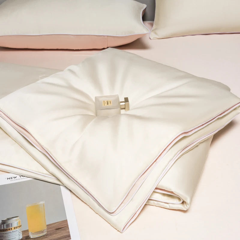 A stack of cream-colored ice silk bedding, including a quilt and pillows, resting on a light pink surface.