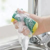 A hand is holding a green and yellow double-sided kitchen scouring pad under running water in a sink.