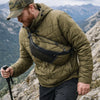 A man wearing a olive green quilted jacket and dark pants is hiking on a rocky mountain trail, carrying an everyday nylon cro