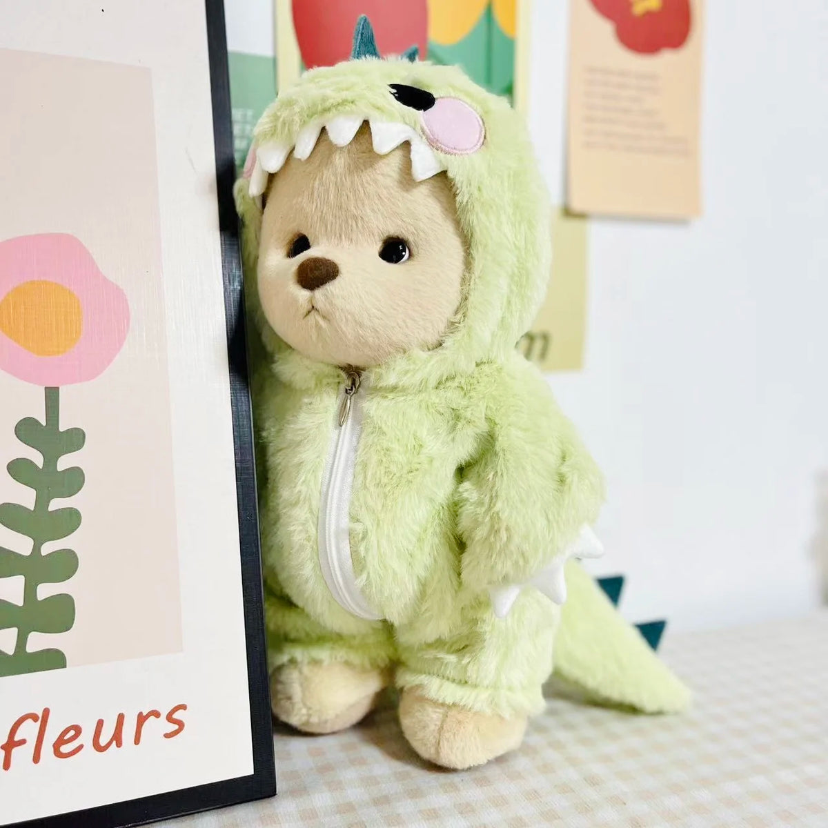 A light green plush teddy bear dressed in a dinosaur jumpsuit outfit.
