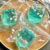 A clear teal glass squeeze toy filled with glitter and small sea creatures, packaged in plastic on a wooden surface.