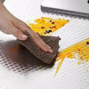 A hand is wiping a dark surface with a brown sponge, leaving yellow oil stains on a white surface.