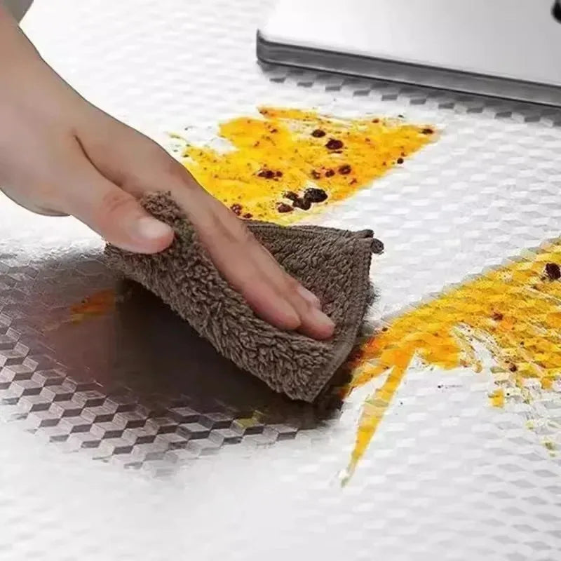 A hand is wiping a dark surface with a brown sponge, leaving yellow oil stains on a white surface.