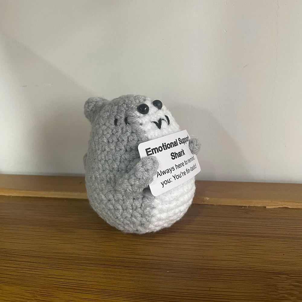 A handmade crochet doll resembling a gray, furry creature with visible teeth and an emotional support tag.