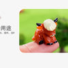 An adorable orange resin Highland cow figurine resting on a person's finger against a blurred green background.