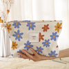 A light-colored floral corduroy makeup bag with blue and orange flowers is being held by a hand.