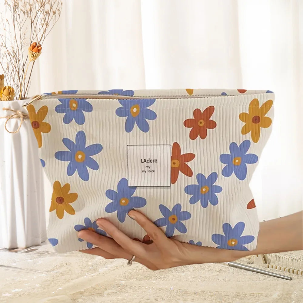 A light-colored floral corduroy makeup bag with blue and orange flowers is being held by a hand.