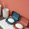 A white leak-proof travel soap box is shown open on a white towel against a coral wall.