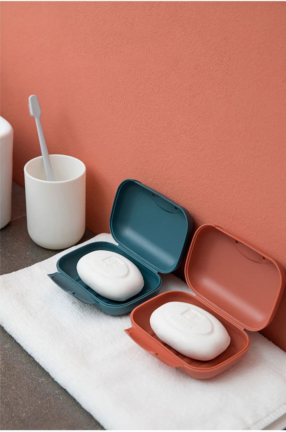 A white leak-proof travel soap box is shown open on a white towel against a coral wall.