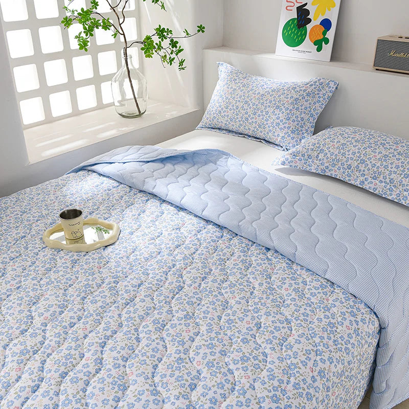 A light blue quilted blanket with a white floral pattern is displayed on a bed next to a window.
