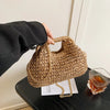 A hand is holding a woven straw chain crossbody handbag on a dark surface.