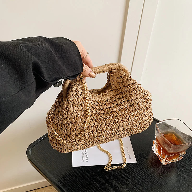 A hand is holding a woven straw chain crossbody handbag on a dark surface.