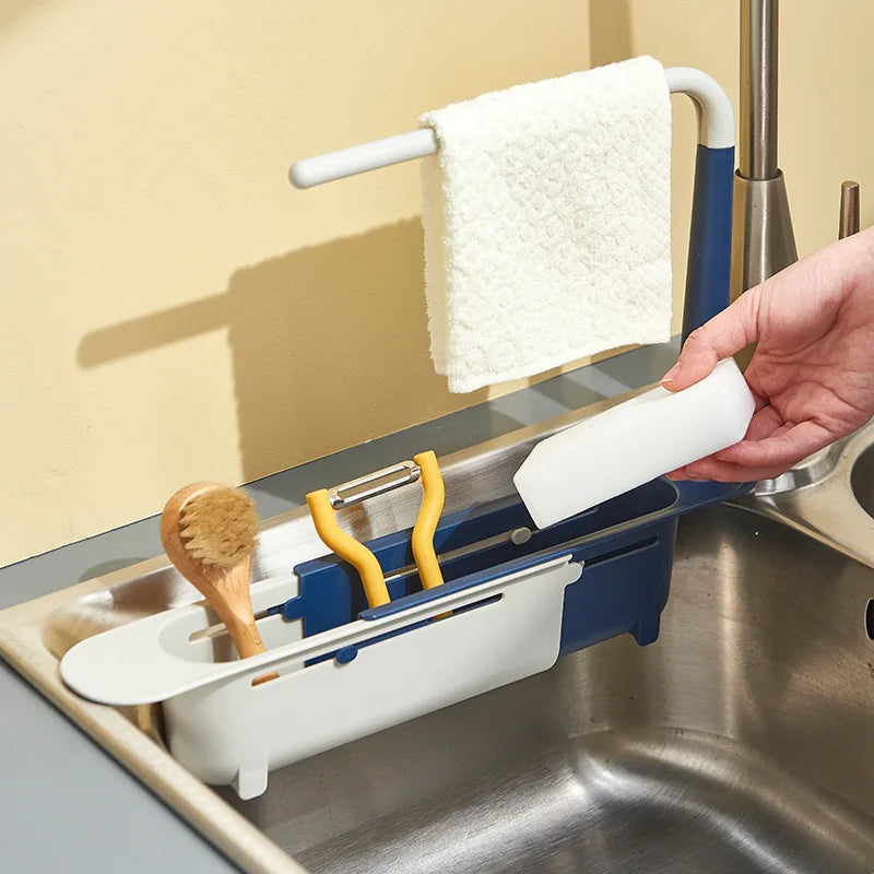 A hand is holding a white sponge hanging from an adjustable sink holder with a drainer basket.