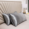 A close-up of a silky smooth satin pillowcase in gray resting on a bed with a padded headboard and a lamp.