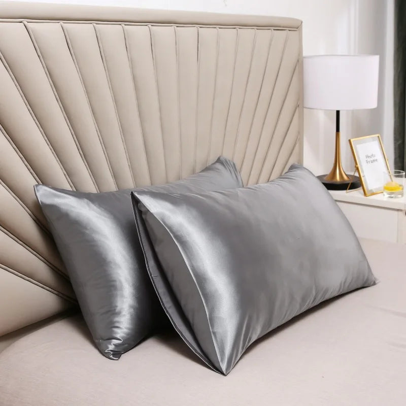 A close-up of a silky smooth satin pillowcase in gray resting on a bed with a padded headboard and a lamp.