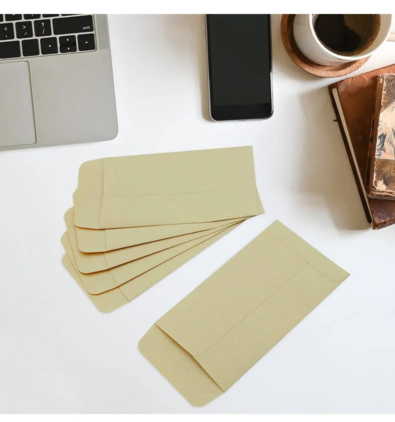 A stack of light tan kraft paper cash envelopes is displayed on a white surface next to a laptop and a smartphone.