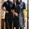 Two women wearing black leather kitchen aprons with pockets and brown straps standing indoors.