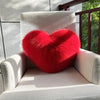 A bright red, fluffy heart-shaped throw pillow resting on a light-colored outdoor chair.
