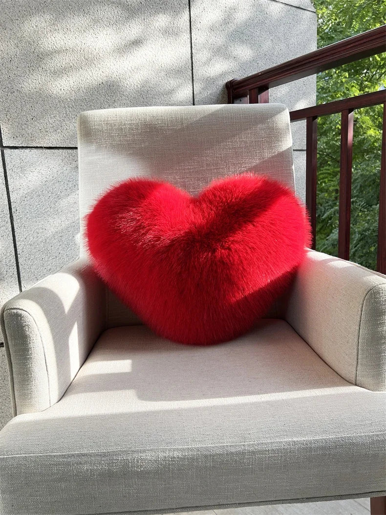 A bright red, fluffy heart-shaped throw pillow resting on a light-colored outdoor chair.
