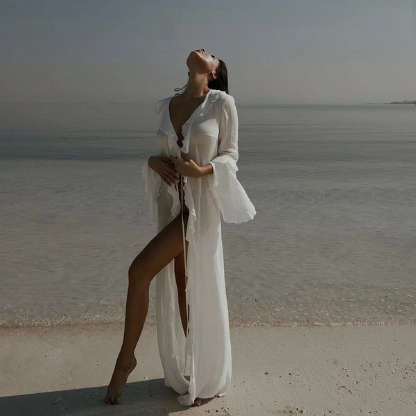 A woman wearing a white chiffon beach cover-up tunic with ruffled sleeves stands looking up toward the sky near the water.
