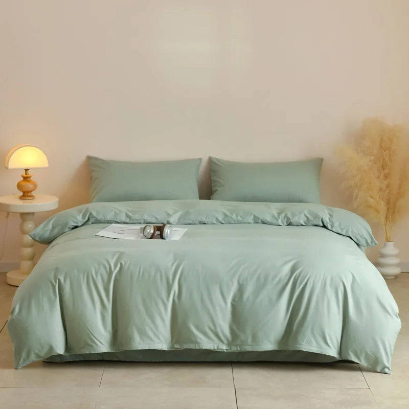 A light sage green quilted duvet cover set with two matching pillowcases on a bed against a white wall.