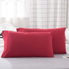 A set of two solid red microfiber pillows resting on a light gray bed with a white checkered curtain in the background.