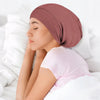 A woman wearing a dusty rose silk-like night cap rests her head on a white pillow.
