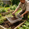A man uses a stainless steel garden soil sieve to fill a plant pot with dark soil in a garden bed.