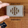 A close-up of a beige embroidered tactical patch featuring a Templar cross design, shown next to a finger and a black hook an