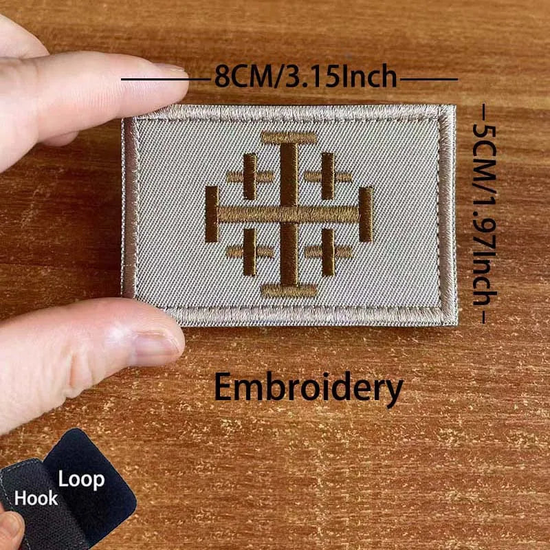 A close-up of a beige embroidered tactical patch featuring a Templar cross design, shown next to a finger and a black hook an
